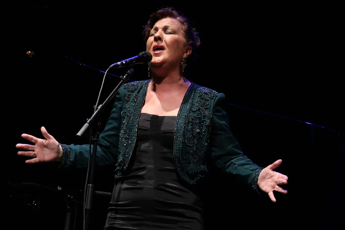 A flamenco singer with open hands, mid-song at a microphone on a dark stage, in a green beaded jacket